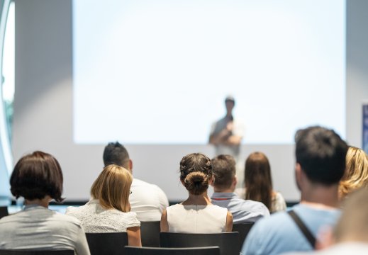 Bilde av mennesker i en sal som hører på et innlegg, bildet er tatt bakfra og uskarpt i bakgrunnen