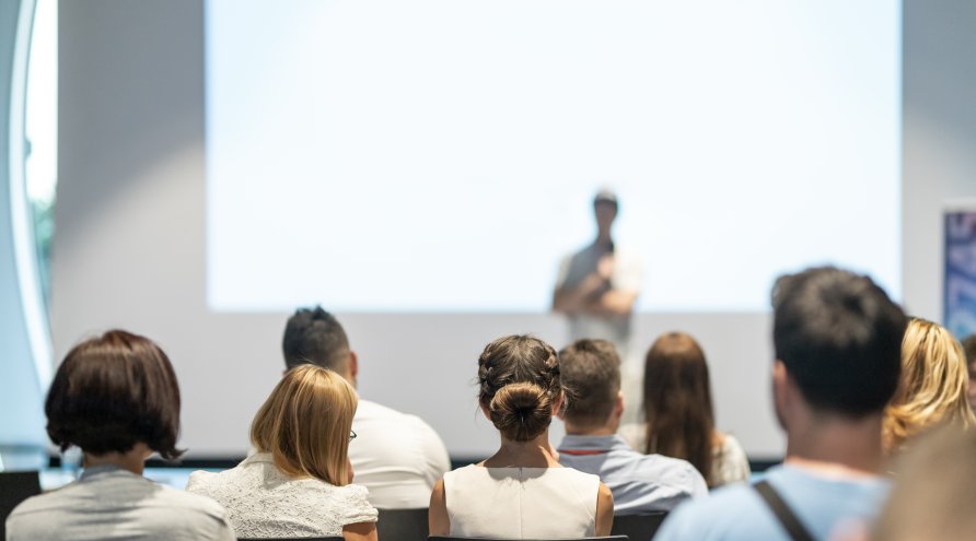 Bilde av mennesker i en sal som hører på et innlegg, bildet er tatt bakfra og uskarpt i bakgrunnen
