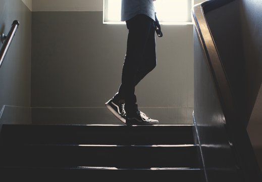 En ung person går oppover i en mørk trappeoppgang kledd i sneakers og bukse, bare nedre del av skjorten vises. Det kommer litt dagslys inn fra vinduet overst på bildet. 
