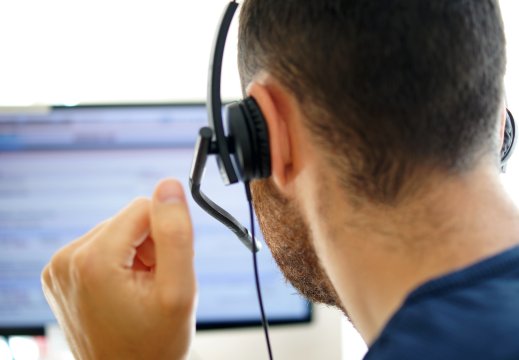 Mannlig bakfra sitter i telefon med headsett og kikke på dataskjerm.