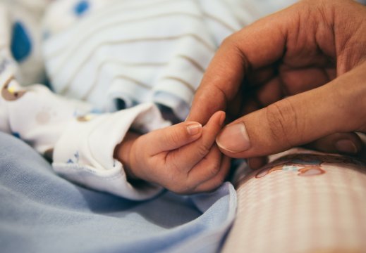 Babyhånd på et blått teppe, tre fingre berøres av en hånd fra en voksen person.