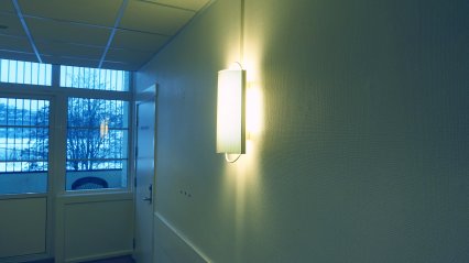 Vegglampe som lyser i en korridor om natten. I enden en dør og en balkong med gitter. Foto.