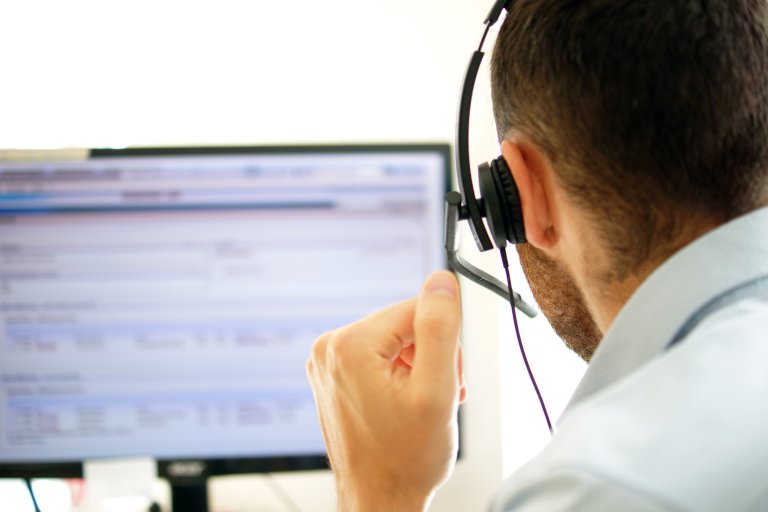 Mann i telefonen med headsett, tatt bakfra, ser inn i dataskjerm, utsnitt av hode/skulde til mann