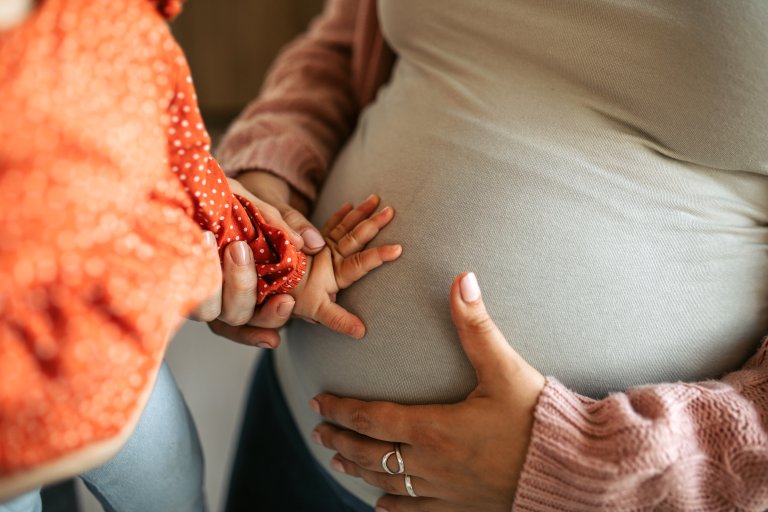 Barnehånd får kjenne på gravid mage, utsnitt av mage, kvinne holder ene hånd på mage, hjelper barnet til å kjenne på magen.