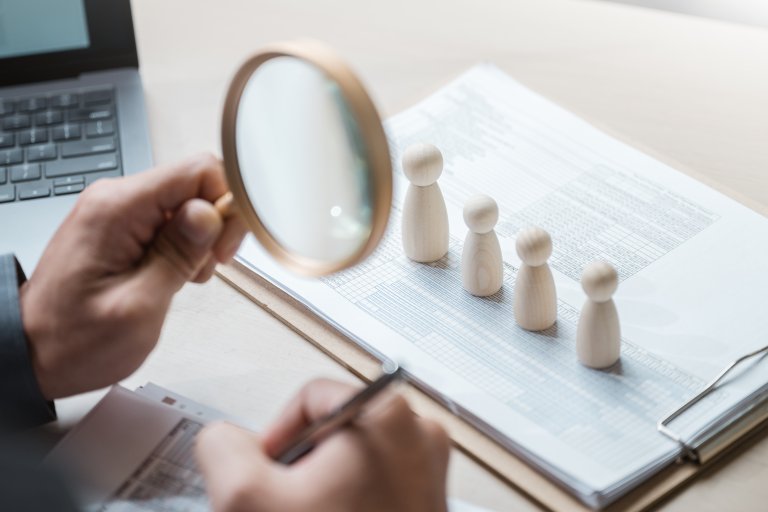 Trefigurer på en rekke_stående på en clipboard med ark. En hånd holder et forstørrelsesglass mot figurer og en penn i andre hånd