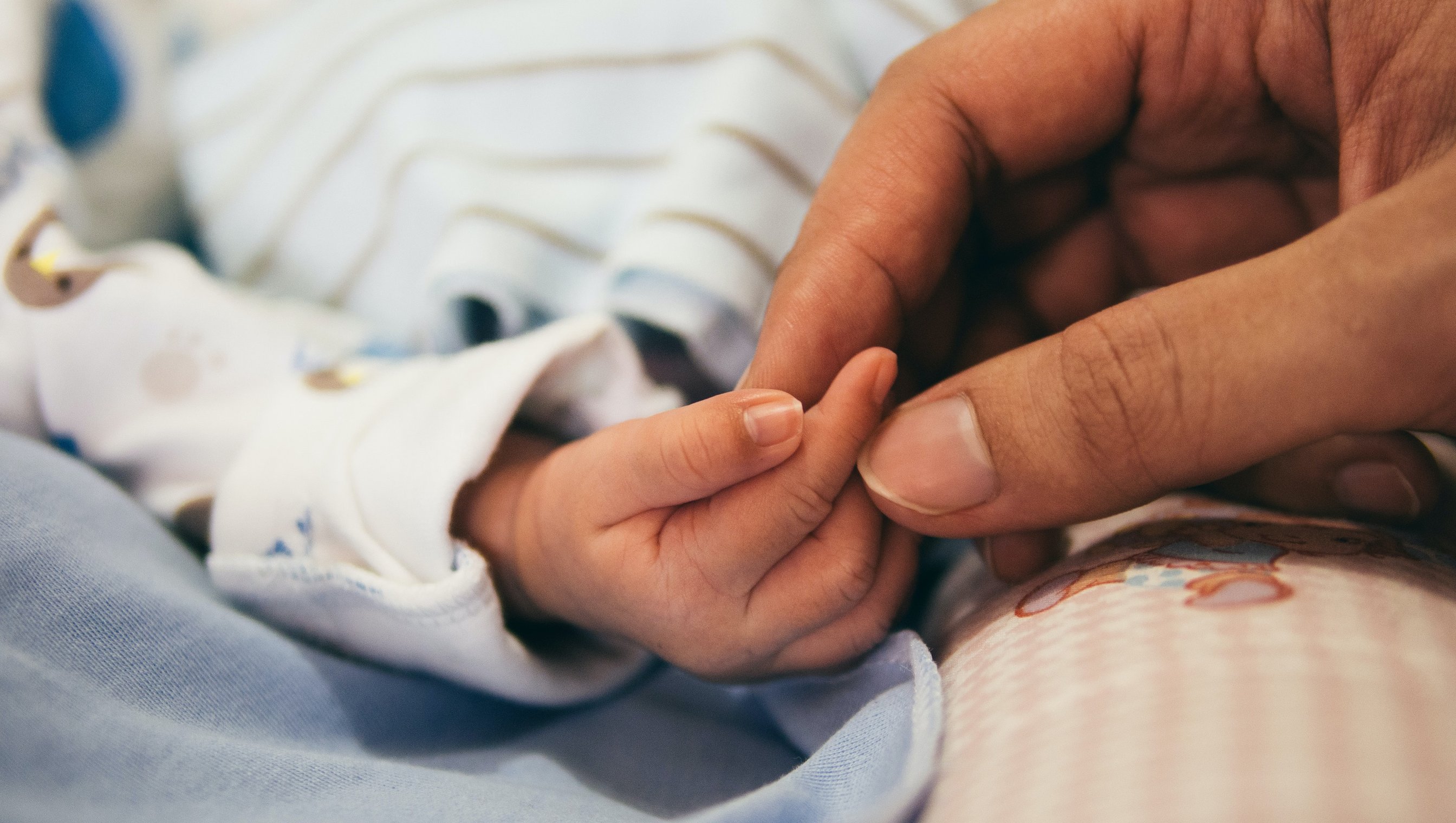 Babyhånd på et blått teppe, tre fingre berøres av en hånd fra en voksen person.